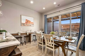 A dining area with a large window, wooden chairs, a table set for two, and a desert landscape painting on the wall. - Ocotillo Springs 53, Private Pool, Spa, Resort Amenities (Santa Clara)