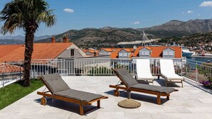 En tagterrasse med liggestole, en palme og udsigt over en kystby med røde tage og bjerge i baggrunden.