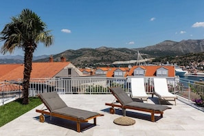 Eine Dachterrasse mit Liegestühlen, einer Palme und Blick auf eine Küstenstadt mit roten Dächern und Bergen im Hintergrund.