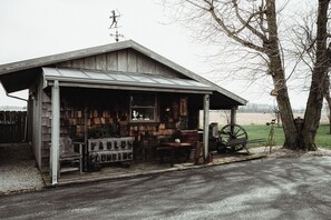 A rustic wooden building with a metal roof, a wooden bench, a wheel, and a tree in the background.