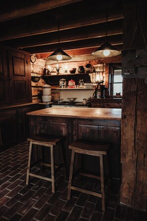 A rustic kitchen with a wooden island, stools, hanging pendant lights, and a brick floor.