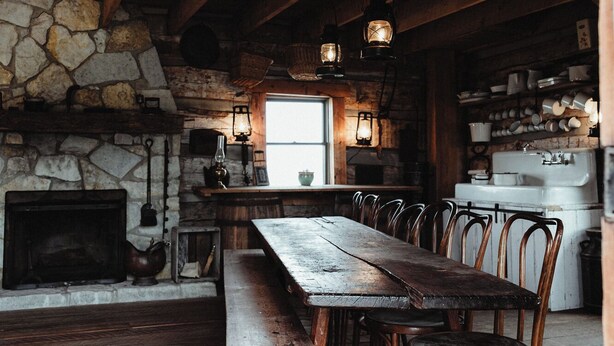 Un intérieur rustique avec des éléments en bois, une cheminée en pierre, des poutres apparentes, une longue table à manger entourée de chaises et des lanternes suspendues.