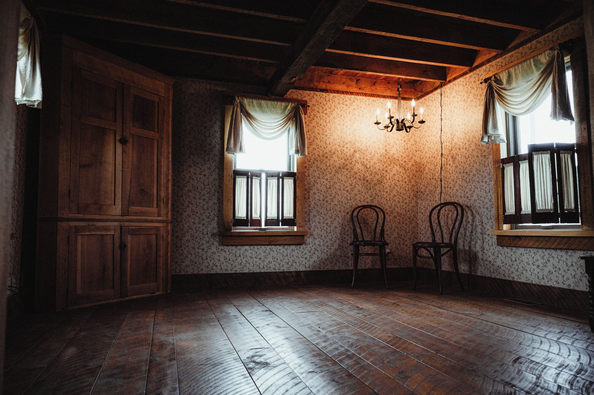 Una habitación con pisos de madera, dos ventanas con cortinas, una araña y un armario de madera.