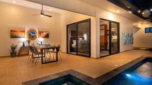 Zona piscina interna moderna con tavolo e sedie, ventilatore a soffitto e un'ampia porta-finestra scorrevole che dà su una camera da letto.