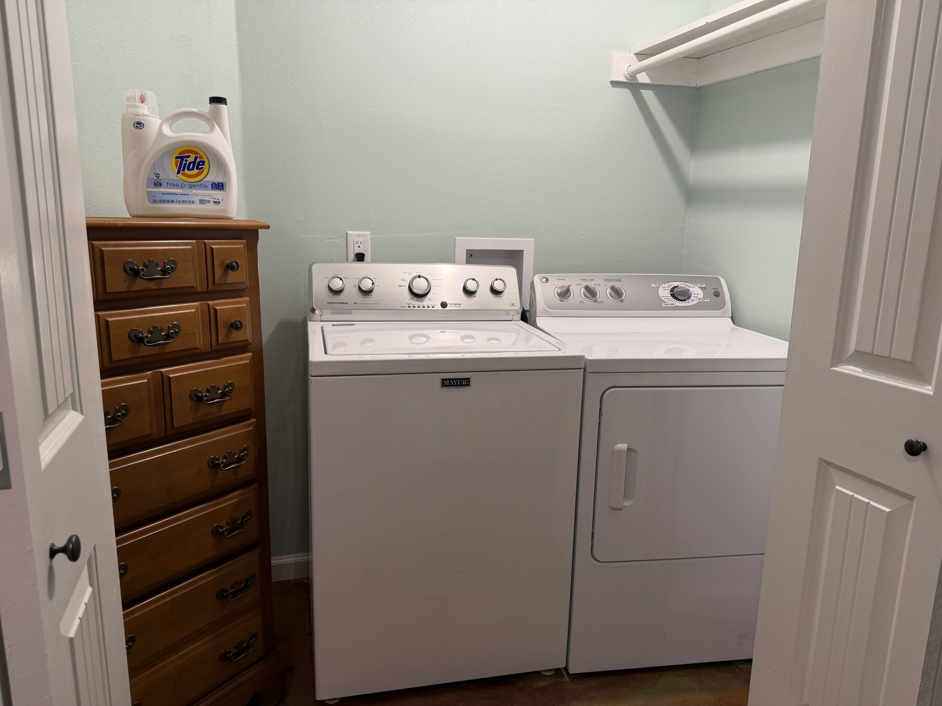 Une buanderie avec un lave-linge et un sèche-linge blancs, une commode en bois avec des tiroirs, et une bouteille de lessive Tide posée sur le dessus.