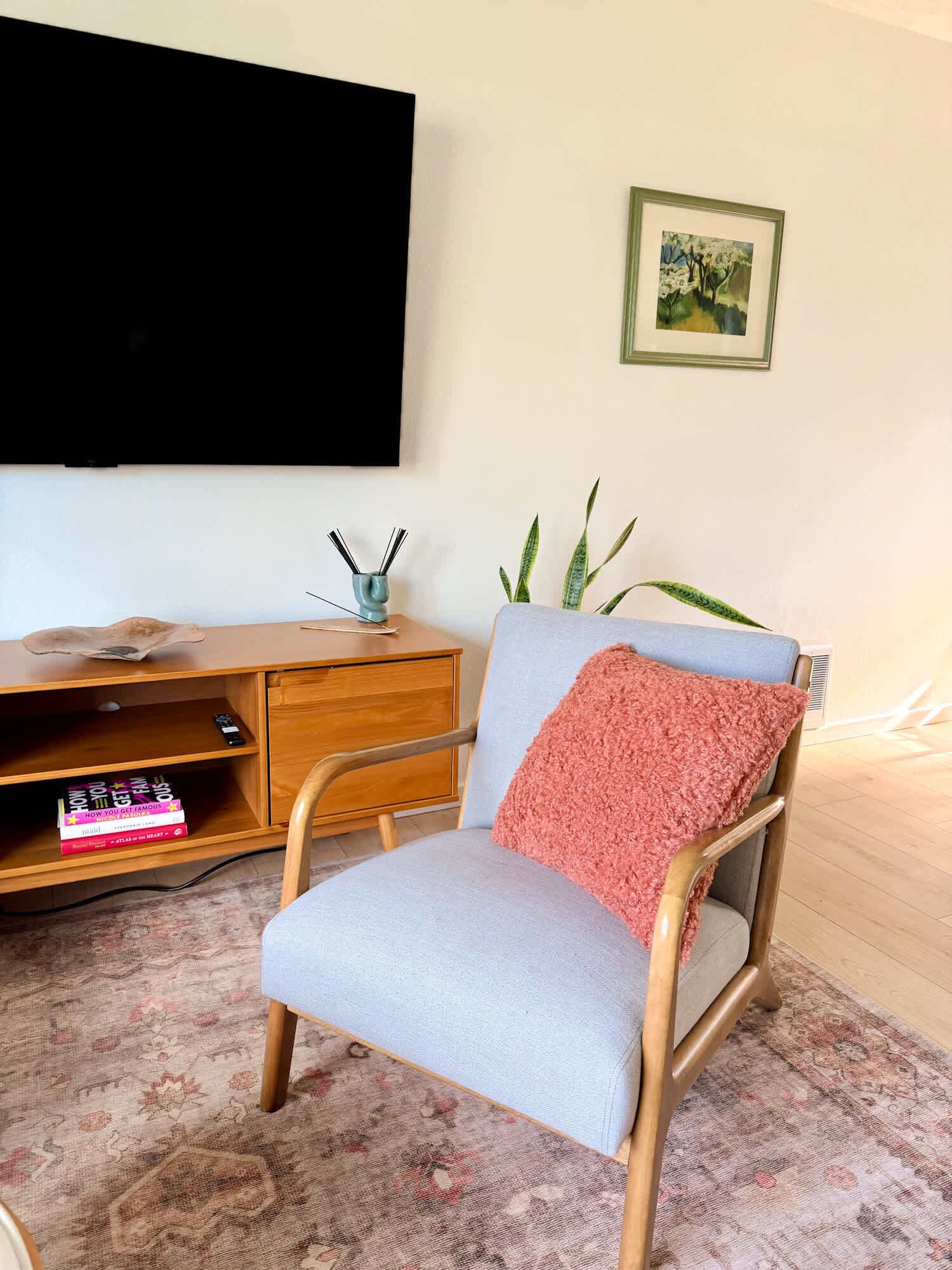 A room with a wooden desk, a chair with a pink throw, a plant, and a framed picture on the wall.