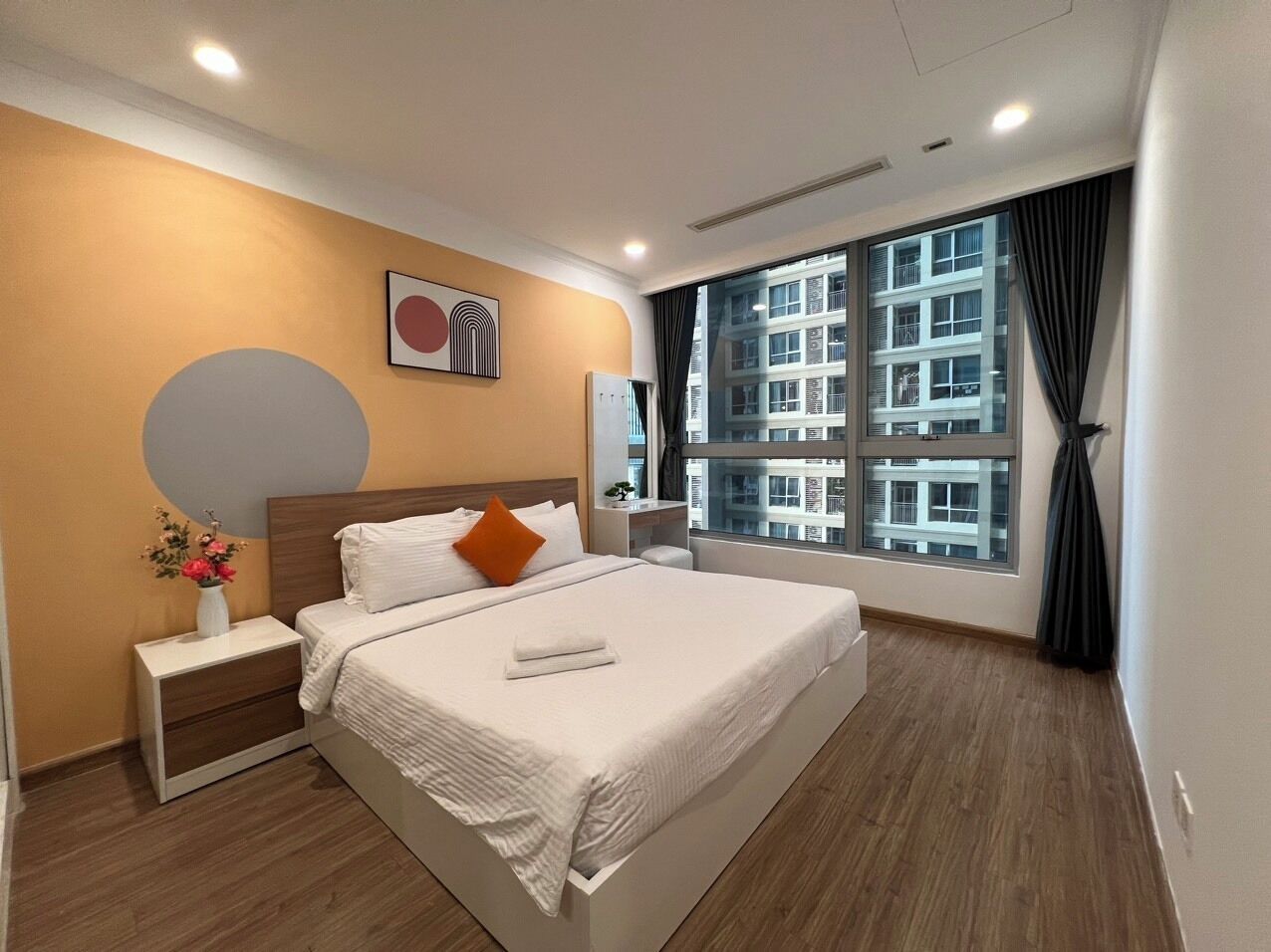 A modern hotel room with a large bed, a bedside table with a vase of flowers, a window with a view of buildings, and a wall with a circular design.