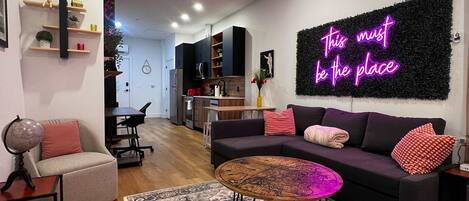 A modern living room with a sofa, coffee table, and a rug. There is a neon sign on the wall and a kitchen in the background.