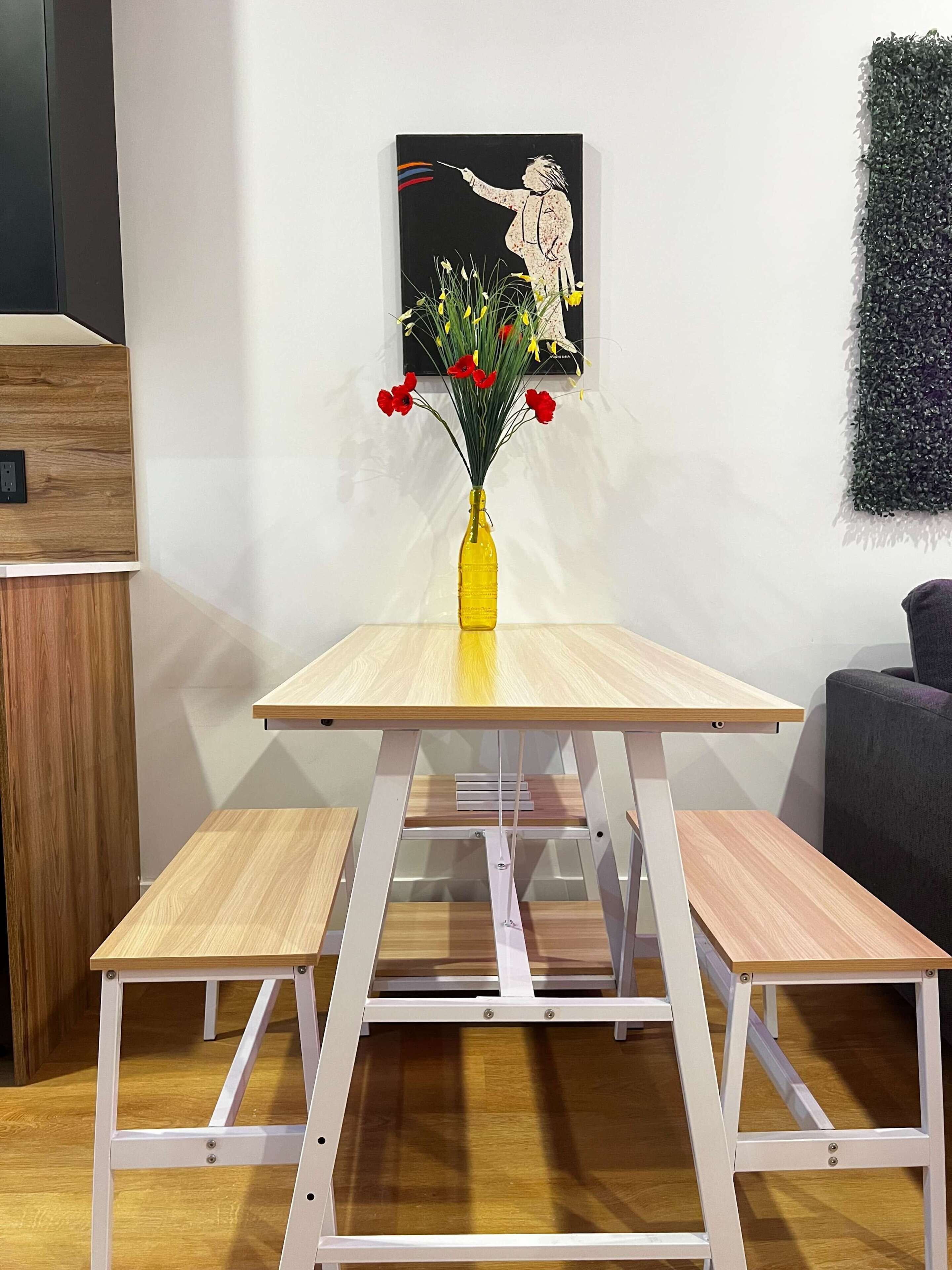 A table with a yellow vase of flowers, a painting on the wall, and a grey couch.