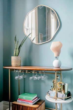 Una habitación con un espejo redondo, una planta en maceta, una lámpara, una mesa con libros y una estantería decorativa.