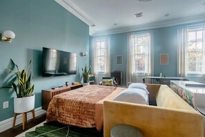 A bedroom with a bed, a desk, a chair, a TV, and two windows.