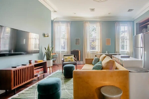 Una sala de estar moderna con un televisor de pantalla plana, un sofá, otomanas, una mesa de centro y una cocina con refrigerador.
