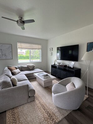 Ein modernes Wohnzimmer mit einer Eckcouch, einem Couchtisch, einem runden Sessel, einem Fernseher und einem Deckenventilator.