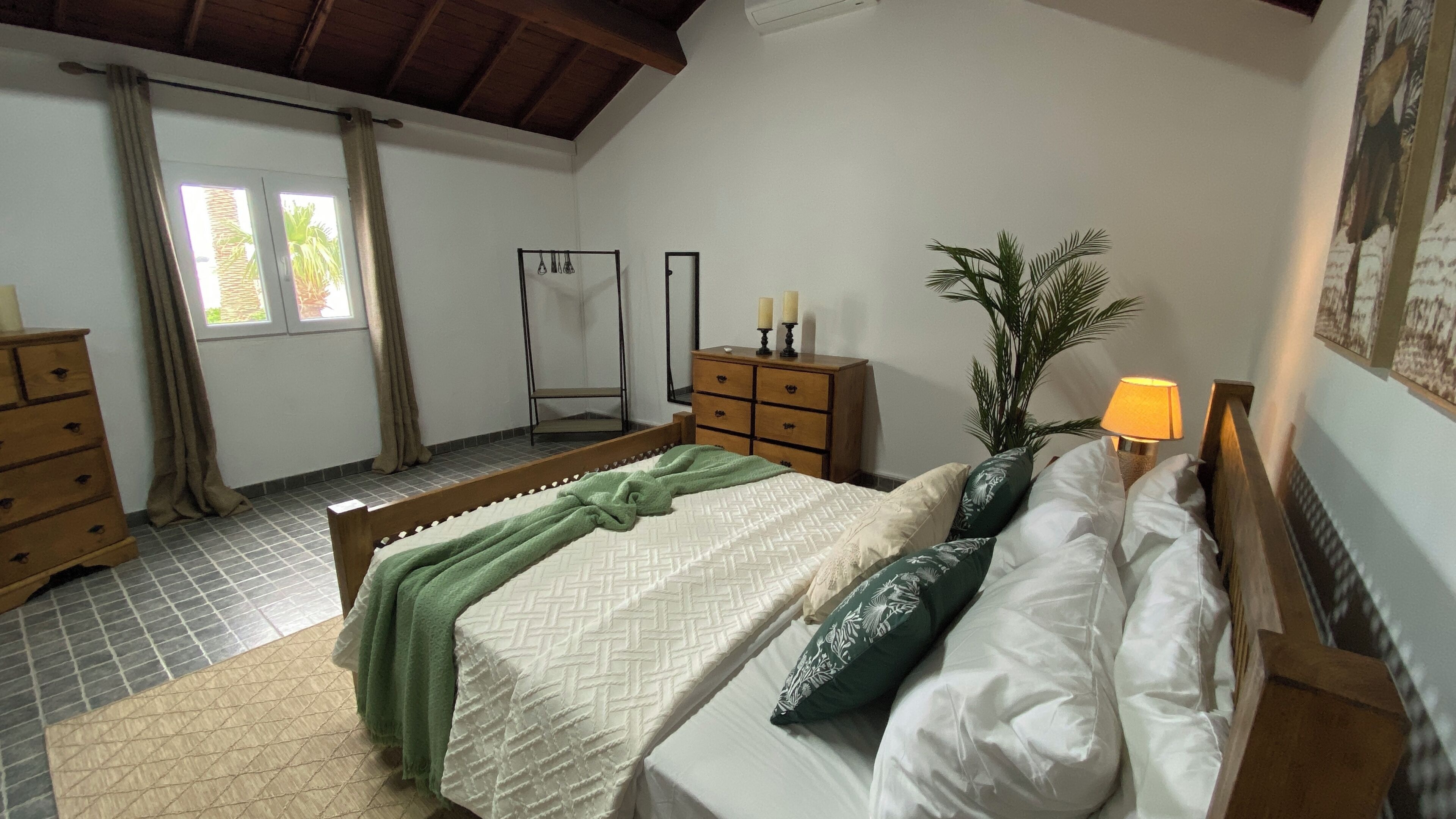 Ein Schlafzimmer mit Bett, Kommode, Nachttisch, Lampe und einem Fenster mit Vorhängen.