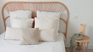 Un lit avec une tête de lit en osier, une literie blanche et une petite table de chevet agrémentée d’une plante.
