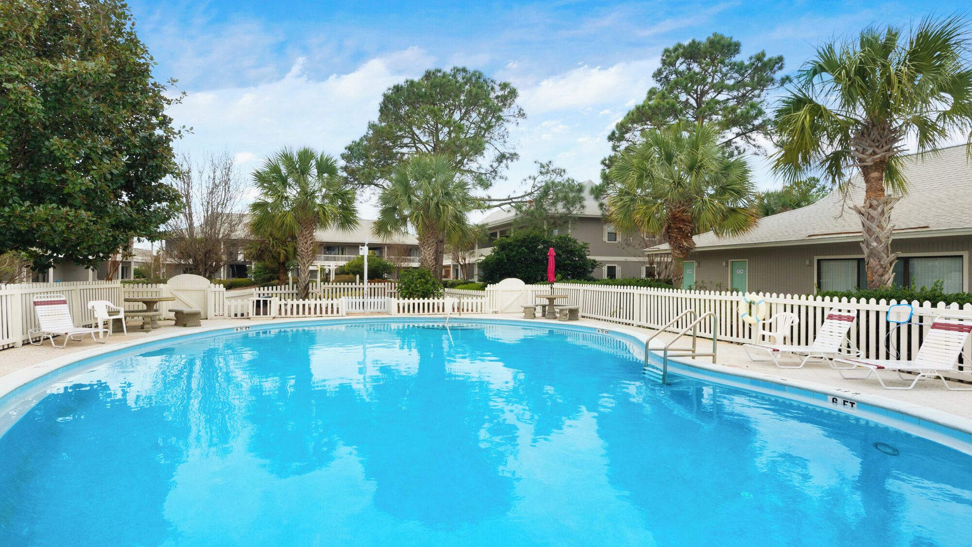 A swimming pool surrounded by a white picket fence, with palm trees and a building in the background.