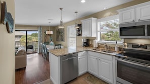 A kitchen with white cabinets, stainless steel appliances, a granite countertop, and a dining area with a view. - Beachwood Villas 10C - 2 Complex Pools, Tennis, Modern Coastal Charm (Santa Rosa Beach)