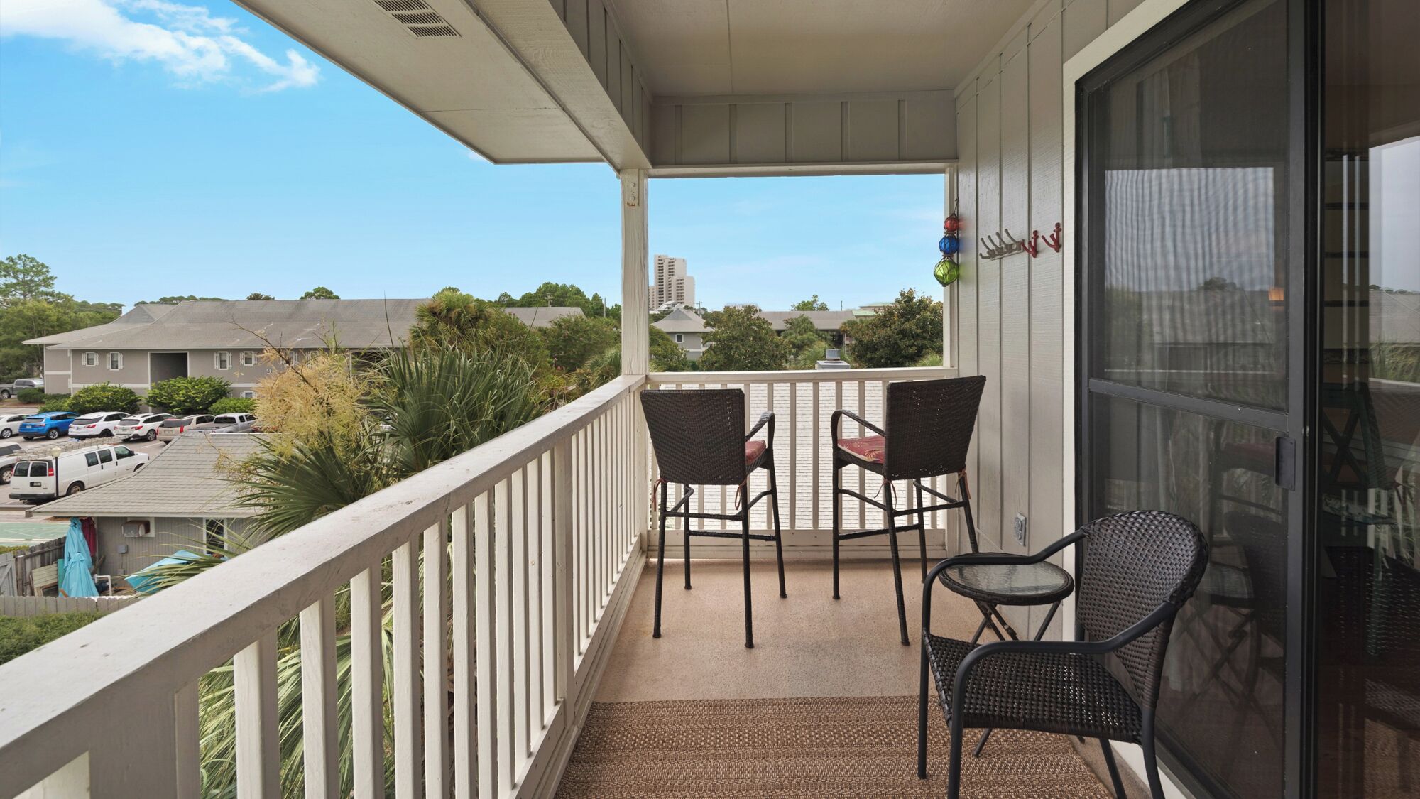 A balcony with two wicker chairs and a small table, overlooking a parking lot and buildings.
