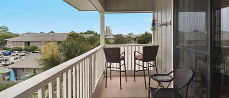 A balcony with two wicker chairs and a small table, overlooking a parking lot and buildings.