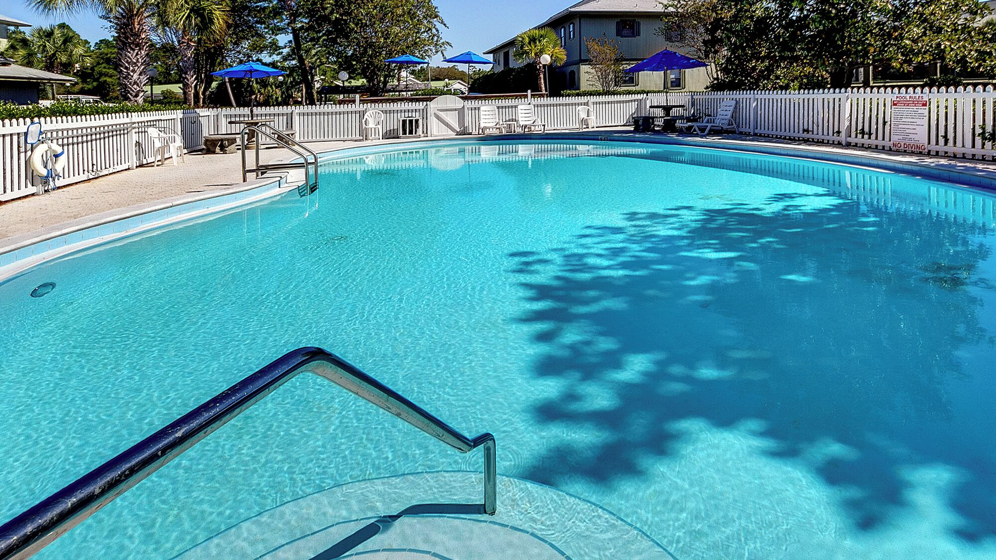 A clear swimming pool with a white fence, blue umbrellas, and a lifeguard chair.