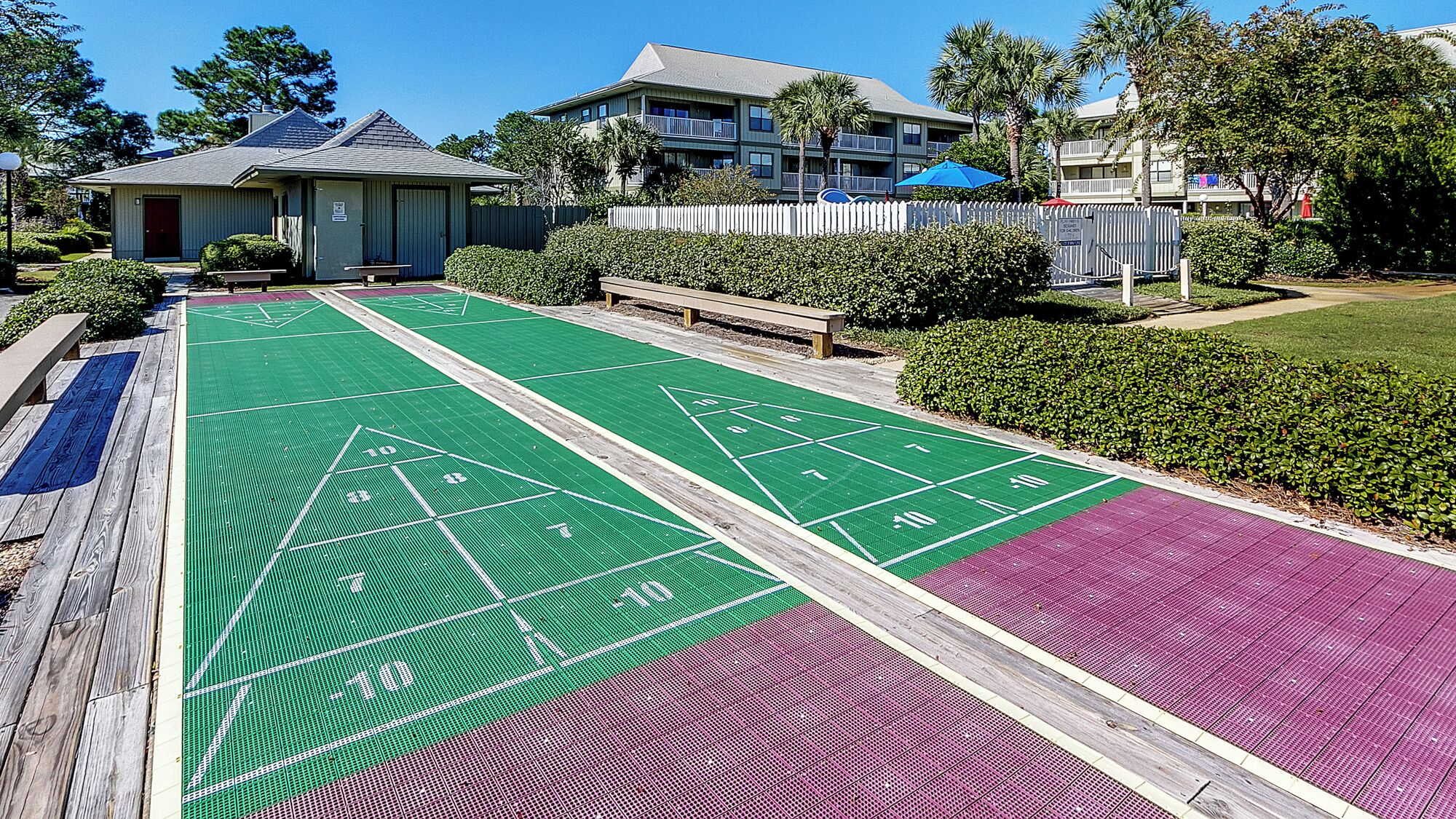 A shuffleboard court with a wooden bench and a small building in the background.