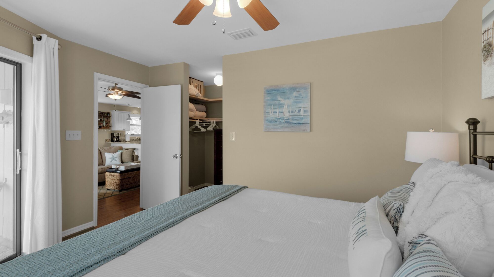 A bedroom with a large bed, a closet with hanging clothes, a painting on the wall, and a ceiling fan.