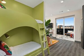 Un lit superposé avec une échelle, une petite table avec une plante et une vue sur la ville à travers une porte vitrée.