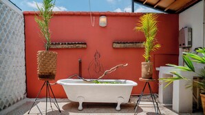 Une salle de bain avec une baignoire blanche, deux plantes en pot, un luminaire fixé au mur et un chauffe-eau.