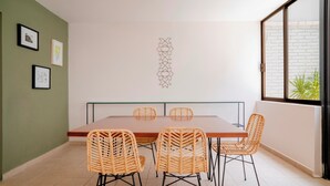 Un coin repas avec une longue table et des chaises en osier, un mur orné de tableaux encadrés et une fenêtre donnant sur un paysage verdoyant.