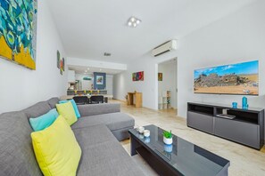 A modern living room with a sectional sofa, a flat-screen TV, and a dining area in the background. - Cozy 1BR/1BA Isla Bonita Getaway Pool&BBQ (Noord)