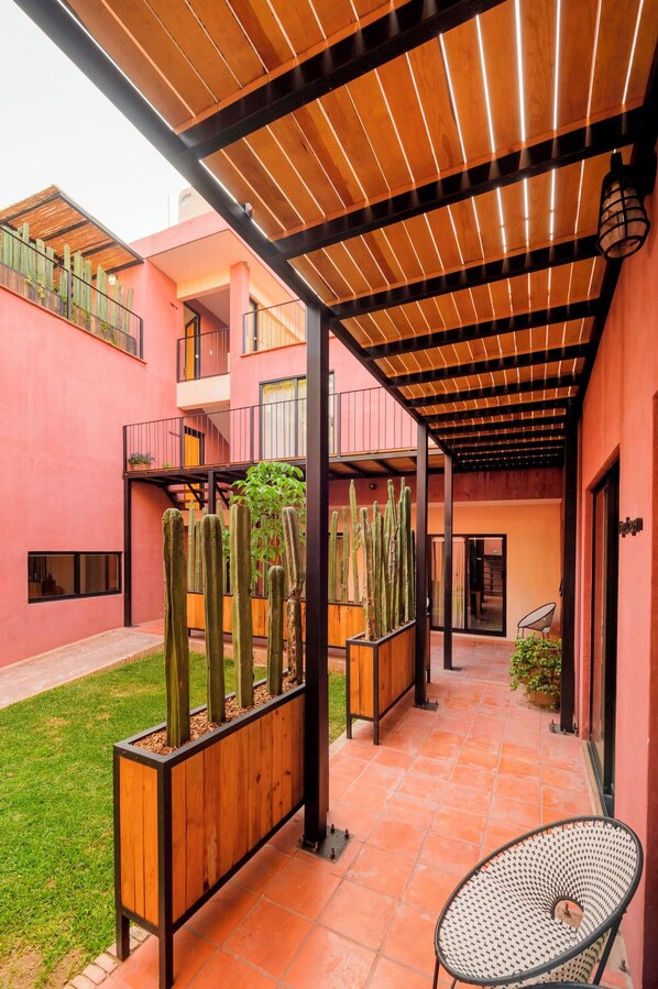 Un espacio al aire libre techado con techo de madera, una maceta con cactus, una silla y un edificio con balcones.