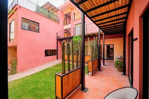 Un patio con piso de baldosas, macetas de madera con cactus, una pérgola de metal y un edificio con paredes rosas y balcones.