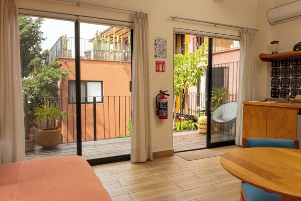 A room with a bed, a wooden table, a chair, a fire extinguisher, and a view of the outside balcony.