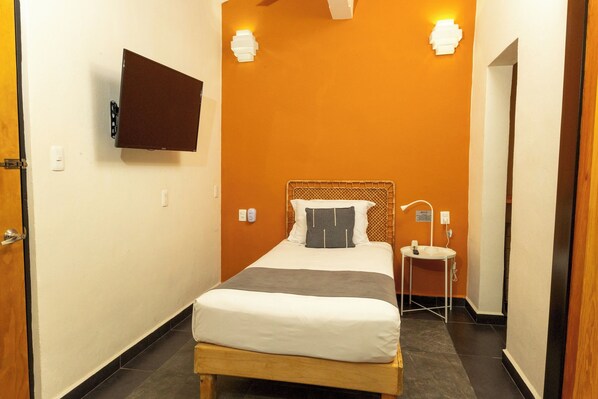 Une chambre d’hôtel avec un lit, un téléviseur fixé au mur, une table de chevet avec une lampe et une petite fenêtre.
