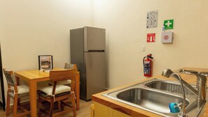 Una cocina compacta con un lavamanos de acero inoxidable, un refrigerador, una mesa de comedor de madera, sillas y un extintor de incendios montado en la pared.