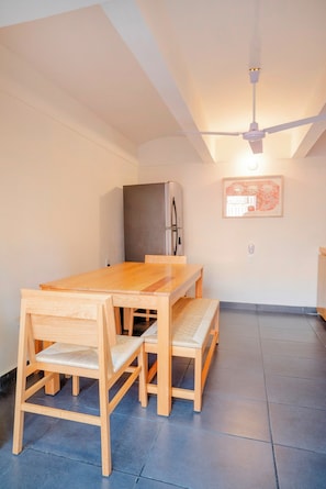 A dining area with a wooden table and chairs, a ceiling fan, a refrigerator, and a framed picture on the wall.