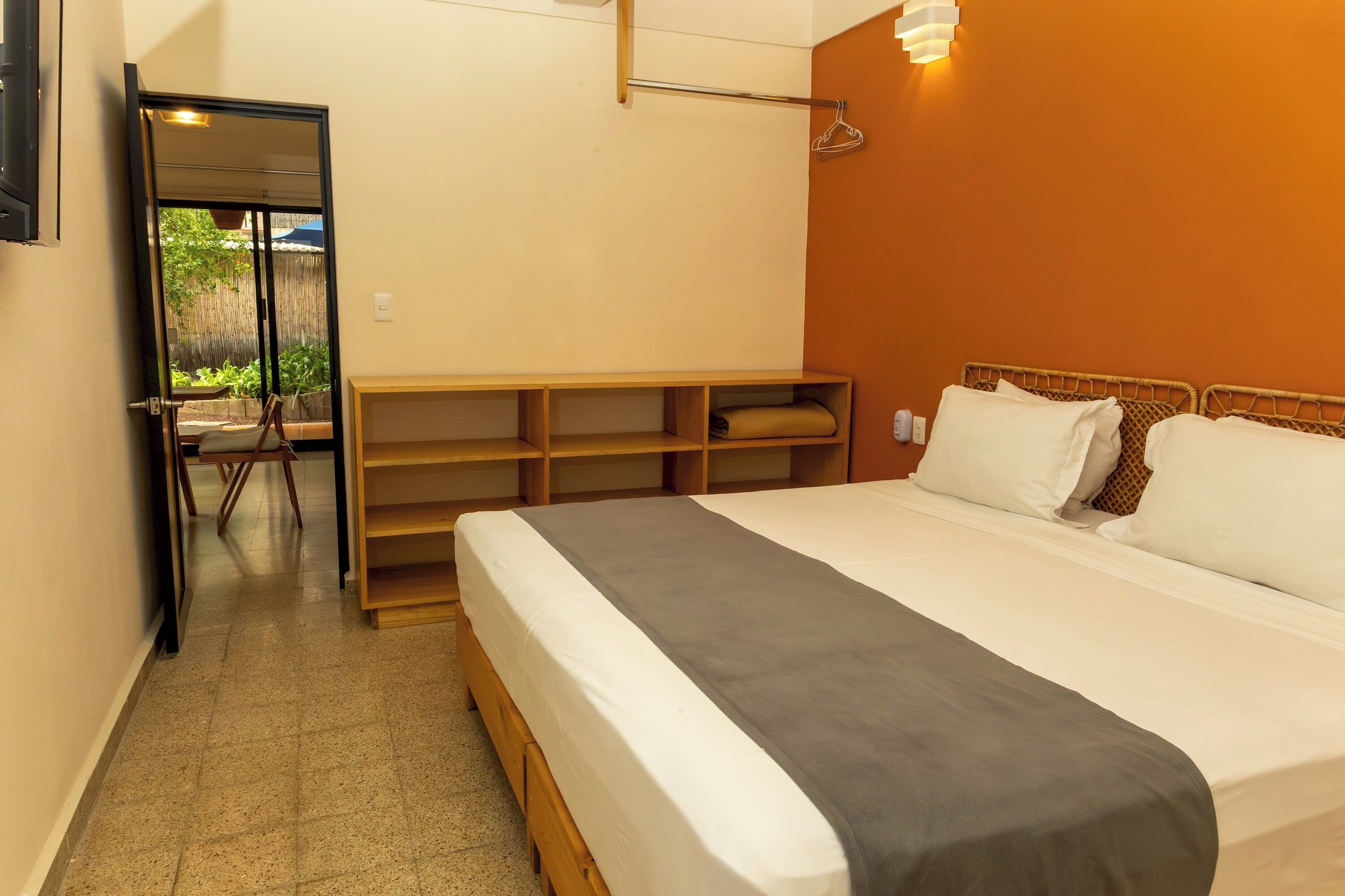 A hotel room with a bed, a wooden bench, a mirror, and a wall-mounted light fixture.