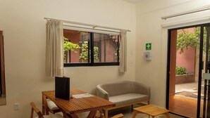 Ein Zimmer mit einem Holztisch, zwei Stühlen, einem Sofa, einem Fenster mit Vorhängen und einer zur Außenseite führenden Tür.