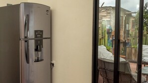 Une pièce de taille modeste, dotée d’un réfrigérateur en acier inoxydable, d’une porte-fenêtre coulissante donnant sur un espace extérieur avec une table et des chaises, et d’un sol carrelé.