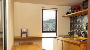 Une cuisine avec une table et des chaises en bois, une fenêtre donnant sur la ville et une étagère remplie d’ustensiles de cuisine.