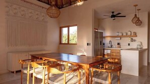 Une salle à manger spacieuse avec un plafond haut et un toit de chaume, une cuisine équipée d’appareils modernes et une grande fenêtre laissant entrer la lumière naturelle.