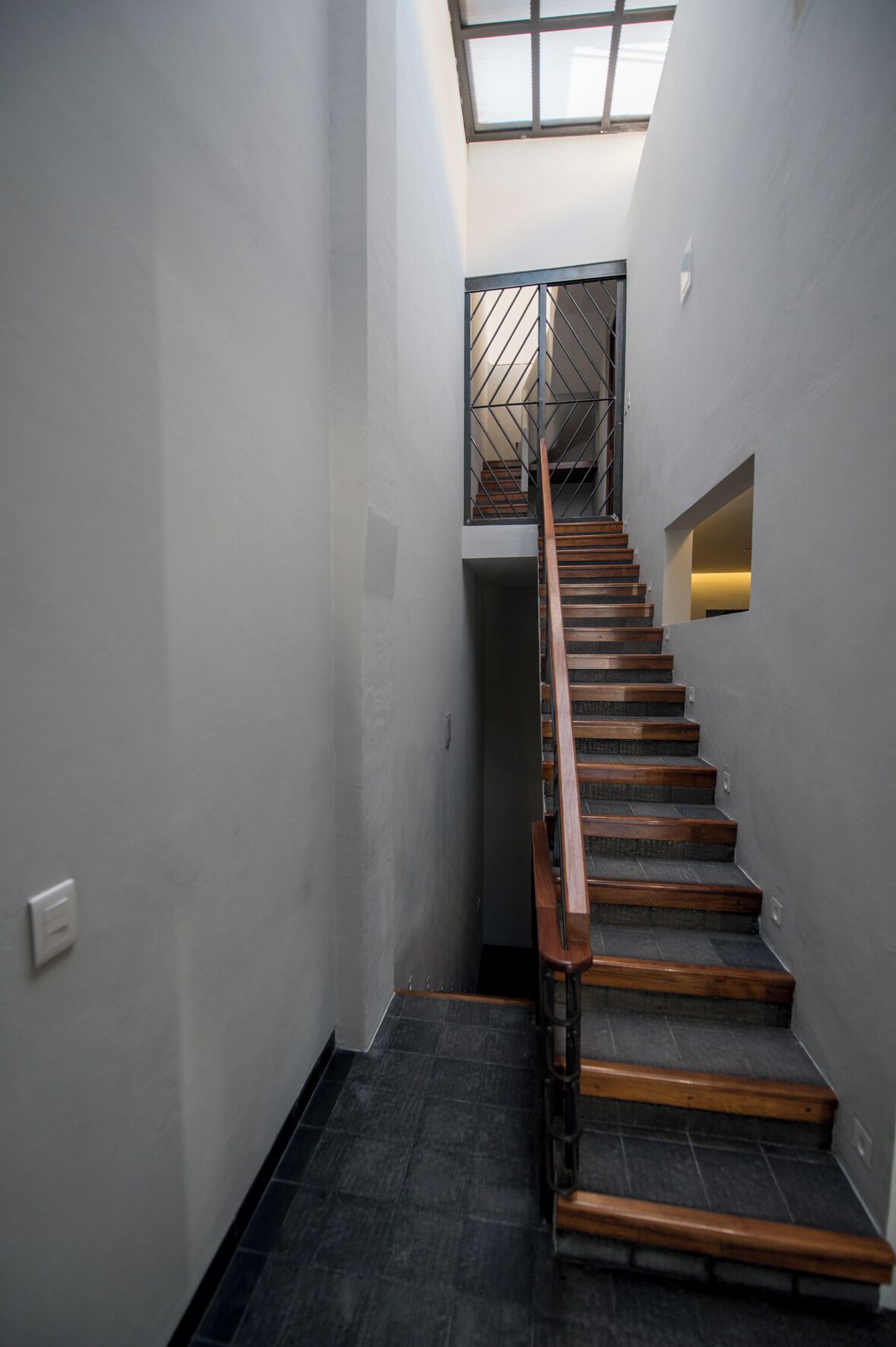 Una escalera estrecha con escalones de madera y barandillas de metal, que lleva a una plataforma con tragaluz.