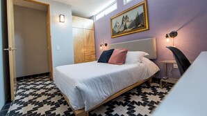 A bedroom with a bed, a wooden wardrobe, a painting on the wall, and a desk with a lamp.