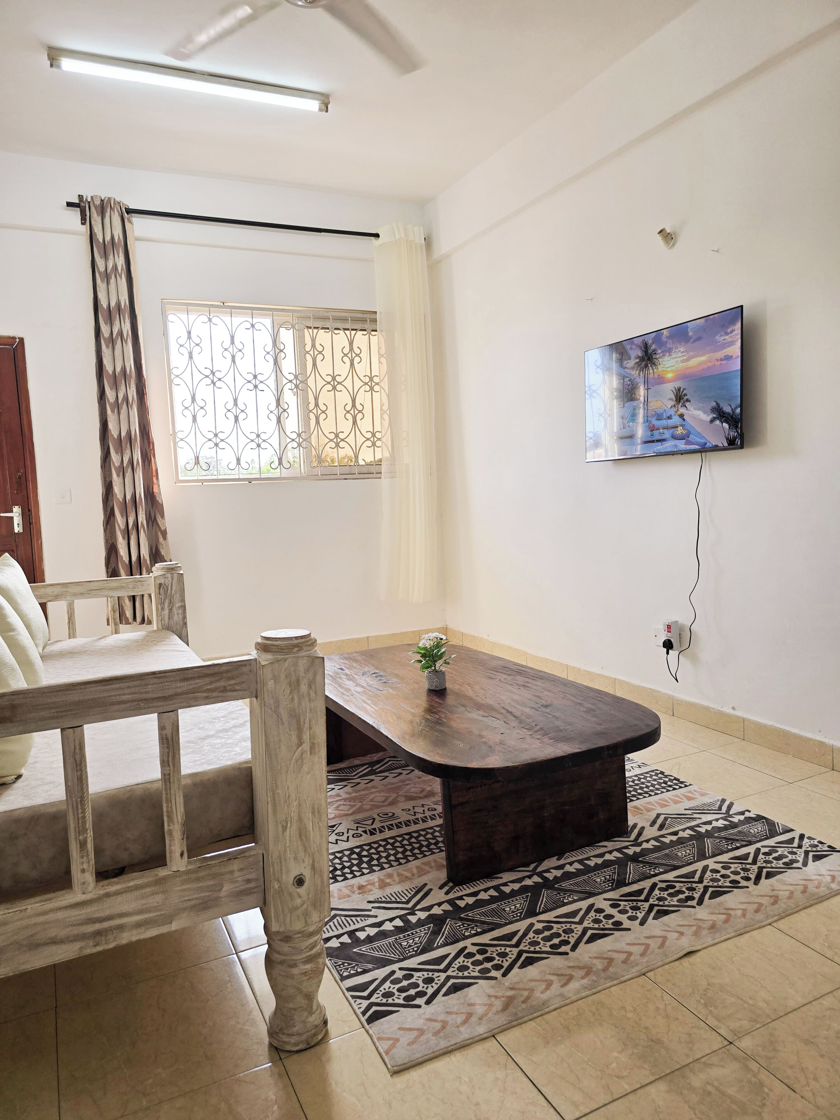 Une pièce avec une table en bois, un tapis à motifs, un téléviseur fixé au mur, une fenêtre ornée de barreaux décoratifs et une porte.