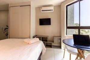 A hotel room with a bed, a TV, a desk with a laptop, a window with a view of buildings, and an air conditioning unit.