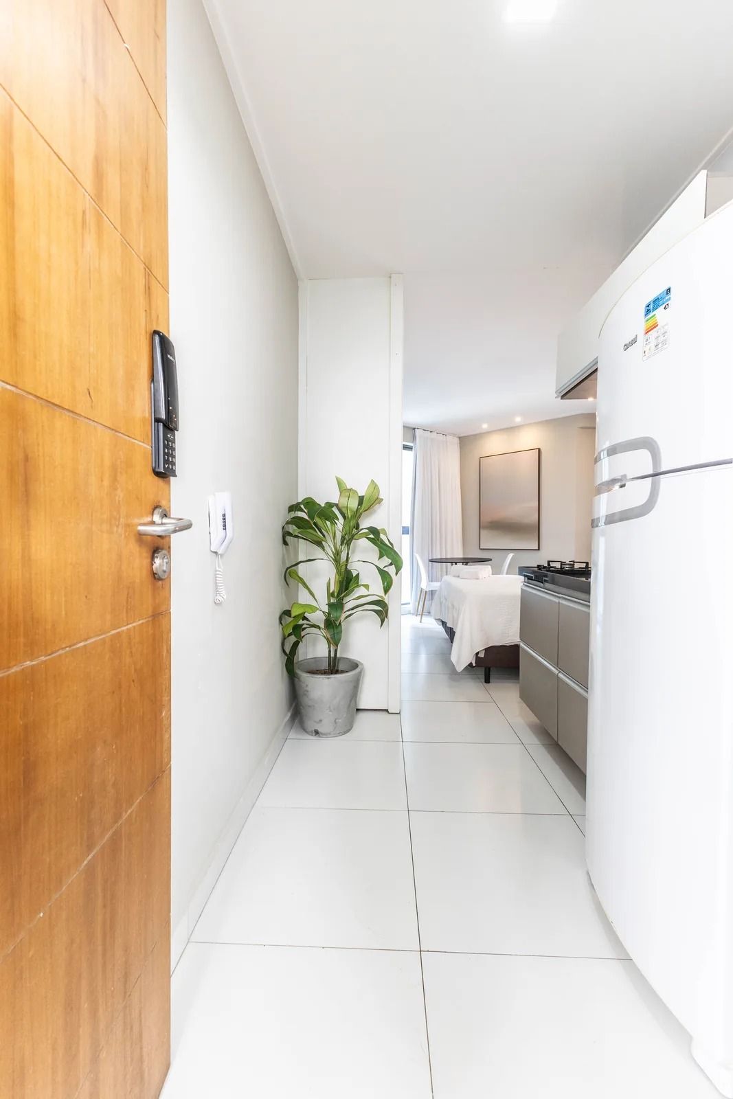 Un pasillo estrecho con una puerta de madera, una planta en maceta, un teléfono y una zona de cocina con refrigerador y estufa.