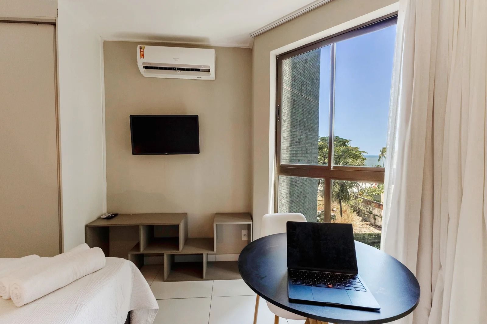A hotel room with a bed, a desk, a laptop, an air conditioner, a TV, and a window with a view of the ocean.