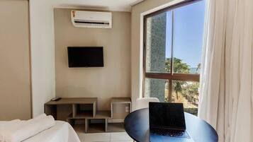 A hotel room with a bed, a desk, a laptop, an air conditioner, a TV, and a window with a view of the ocean.
