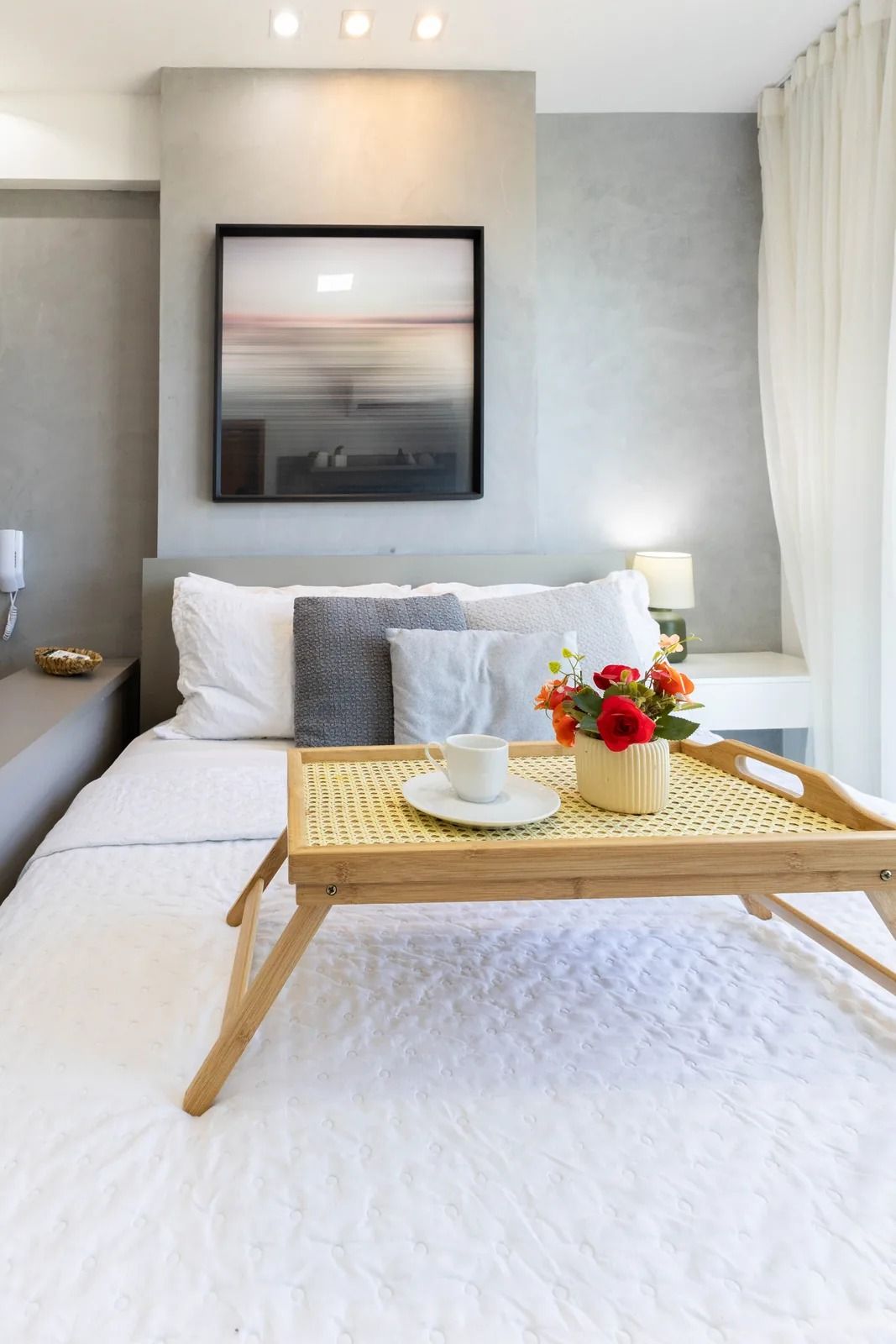 Habitación de hotel con cama, mesita de noche, una lámpara, un cuadro en la pared, una ventana con cortinas y una bandeja con una taza y flores.
