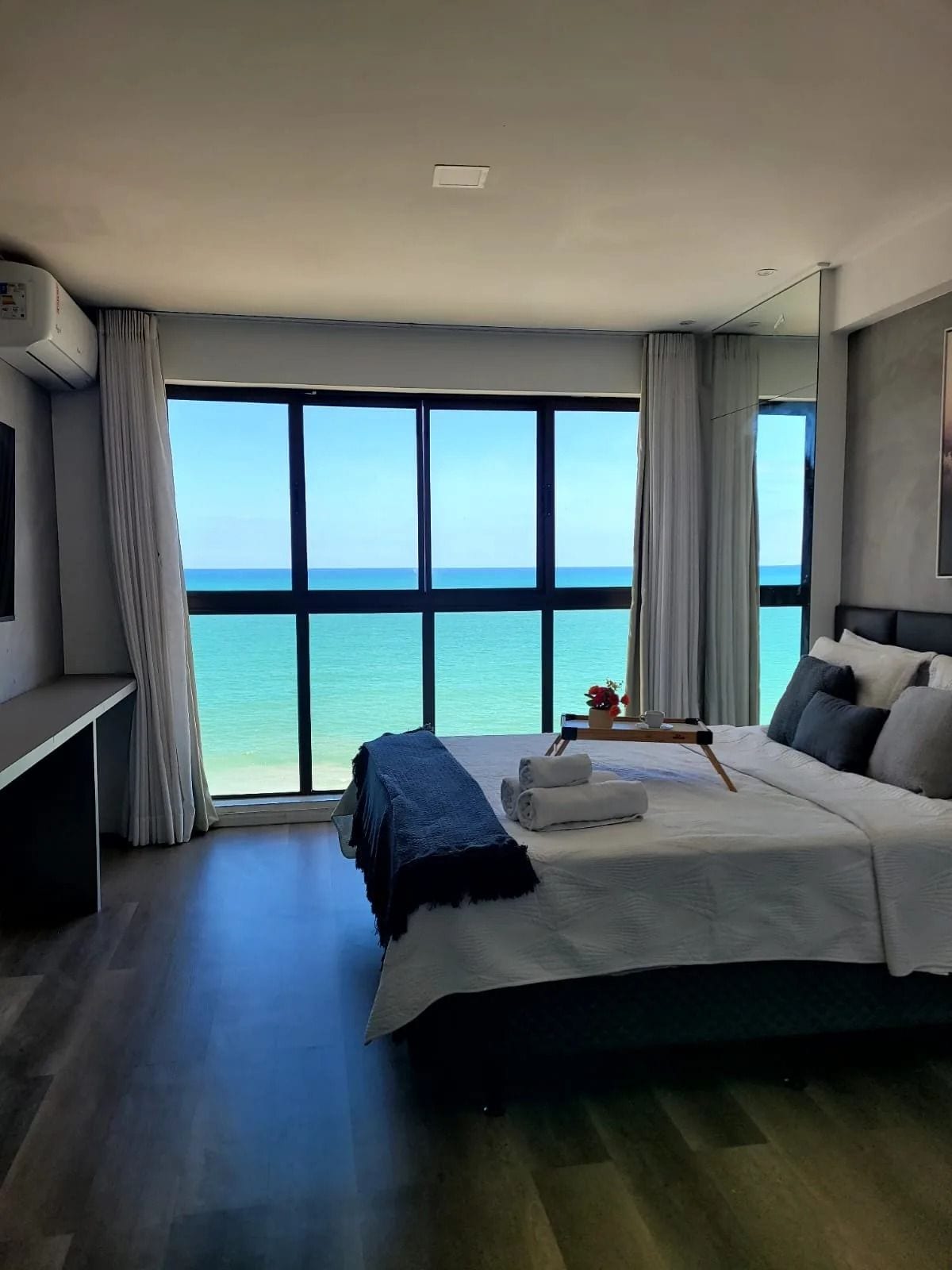 A hotel room with a large window offering a view of the sea, a bed with pillows and a blanket, a bedside table with a tray, and an air conditioning unit.